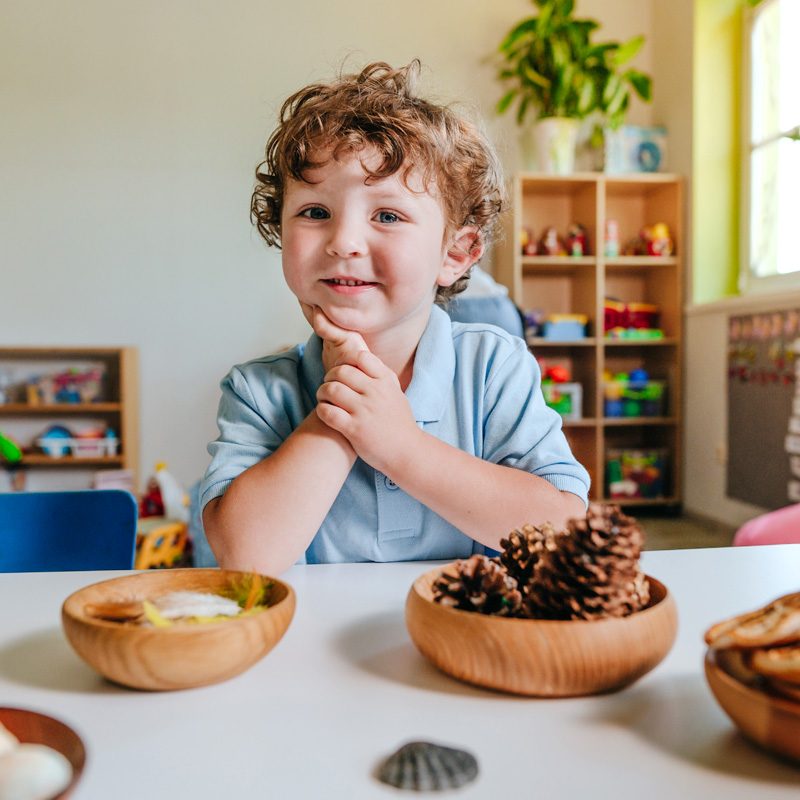 A Pre-Primary child smiling beside bowls of pine cones and shells