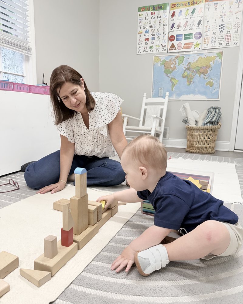 A Montessori guide sitting on the floor beside a toddler building with wooden blocks at AIA Montessori